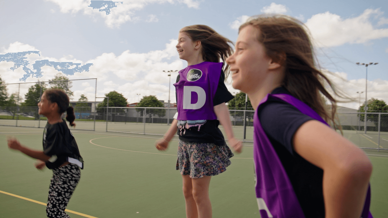 Three girls wearing purple bibs with a “D” on them are smiling and jumping outdoors on a netball court, with a partly cloudy sky and trees in the background.